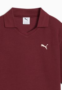 Polo shirt in marrone con un tessuto testurizzato, caratterizzato da un colletto a V e un logo Puma bianco sul lato sinistro del petto.