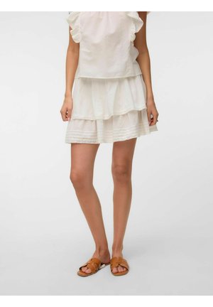 Femme portant une mini-jupe blanche à volants, un top sans manches blanc à volants, et des sandales marron clair enfilables, debout devant un fond clair uni.