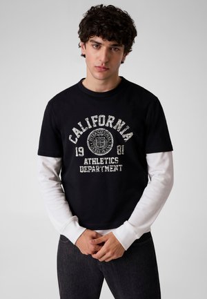 Jeune homme aux cheveux foncés et bouclés porte un t-shirt noir avec l'inscription « "California 1981 Athletics Department" » sur une chemise blanche à manches longues.