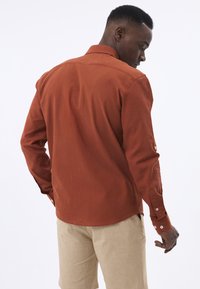 Camisa de botones de color óxido con mangas largas, cuello y un corte relajado. La parte posterior presenta una textura suave y costuras sutiles.