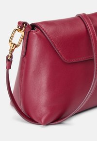 Bolsa de couro vermelho com forma triangular, apresentando um fecho dobrável, ferragens em metal dourado e uma alça destacável. Textura suave.