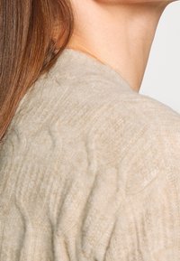 Beige knit sweater with a textured pattern. The fabric appears soft and lightweight, draping smoothly over the shoulder.