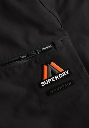 Czarna tkanina z poziomym zamkiem po lewej stronie oraz trójkątną łatką z logo Superdry Mountain w kolorze pomarańczowym, szarym i białym po prawej stronie.