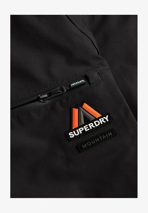 Czarna tkanina z poziomym zamkiem po lewej stronie oraz trójkątną łatką z logo Superdry Mountain w kolorze pomarańczowym, szarym i białym po prawej stronie.