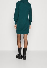 Robe tricotée turquoise avec un col montant, des manches ballon texturées et des poignets côtelés. Associée à des bottines noires à semelle épaisse.