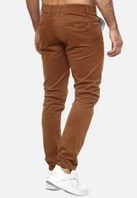 Brązowe dopasowane chinosy o gładkiej fakturze, zwężane nogawki i standardowe tylne kieszenie. Połączone z białymi butami sportowymi.