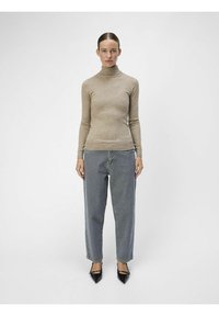 Beżowy sweter z golfem, dopasowany, w połączeniu z luźnymi szarymi dżinsami. Modelka ma na sobie czarne, spiczaste szpilki. Prosty, minimalistyczny design.