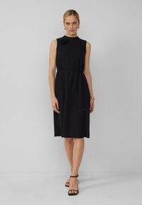 Robe noire sans manches en tissu texturé, encolure ronde, lien sur le côté et accent floral à l'épaule. Le modèle porte des talons hauts noirs à lanières.