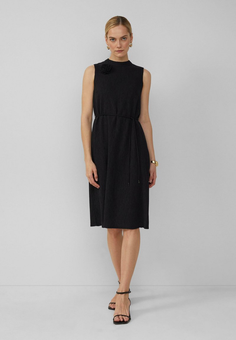 Robe noire sans manches en tissu texturé, encolure ronde, lien sur le côté et accent floral à l'épaule. Le modèle porte des talons hauts noirs à lanières.