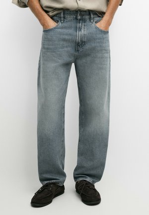 Homme portant un jean ample bleu clair et des chaussures à lacets marron foncé, les mains dans les poches, sur un fond clair uni.