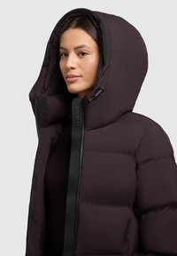 Chaqueta tipo plumón en color púrpura oscuro con cuello alto y capucha de gran tamaño. Cuenta con cremallera negra y tela texturizada para el aislamiento.