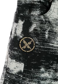 Tejido de patrones en blanco y negro con una superficie texturizada; presenta un logotipo redondo bordado en dorado en un lado.