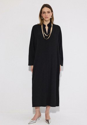 Žena v dlhých čiernych šatách s dlhými rukávmi, vrstvenými náhrdelníkmi zo zlatých mincí, zlatými náušnicami v podobe kruhov a striebornými špicatými podpätkami stojaca pri bielej stene.