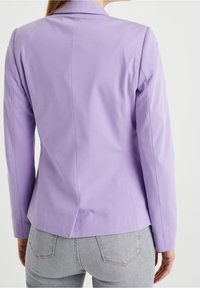 Žena v přiléhavém lila blejzru s dlouhými rukávy a světle šedých džínách, zachycena zezadu na bílém pozadí.