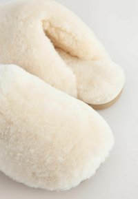 Šviesiai smėliniai pūkuoti šlepetės su minkšta, pūkuota tekstūra. Apskritimo formos, paminkštintos patogumui, turi lygų paviršių.