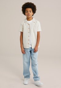 Camicia a maniche corte di colore beige chiaro a righe, con bottoni in legno e taschino sul petto, abbinata a jeans azzurri chiari e sneaker bianche.
