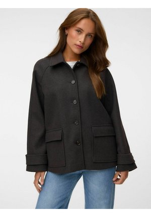 Femme portant une veste en laine sombre boutonnée avec de grandes poches avant et un jean bleu clair sur un fond uni.