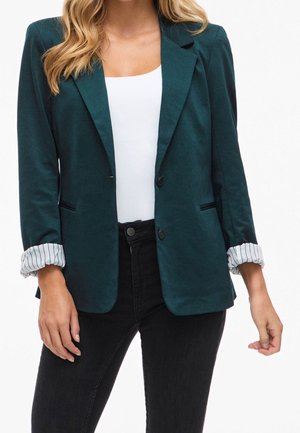 Frau trägt dunkelgrünes Sakko mit hochgekrempelten gestreiften Manschetten, weißes eng anliegendes Oberteil und schwarze hoch taillierte Hose vor weißem Hintergrund.