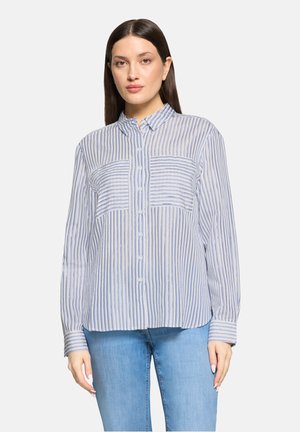 Vrouw draagt een blauw-wit gestreept overhemd met lange mouwen en lichtblauwe spijkerbroek, staand tegen een effen achtergrond.