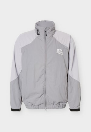 Giacca grigio chiaro con zip, colletto alto, polsini elastici, cinturini neri ai polsi e logo bianco sul petto sinistro.