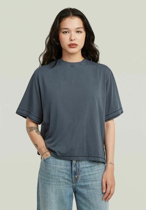 Giovane donna con capelli lunghi e scuri, che indossa una maglietta grigio scuro larga e jeans azzurri, in piedi con una mano in tasca.