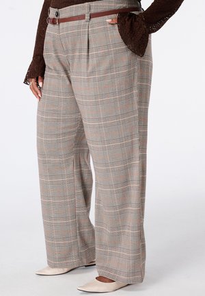Persoon draagt beige geruite wijde broek, bruine riem, bruine getextureerde lange mouwen en crèmekleurige puntige platte schoenen, staand met één hand in de zak.