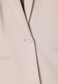 Beige bleiseri, jossa on yksi pyöreä nappikiinnitys, huppureunat ja rakenteelliset hartiat. Sileä pinta, jossa on huomaamattomia ompeluyksityiskohtia.