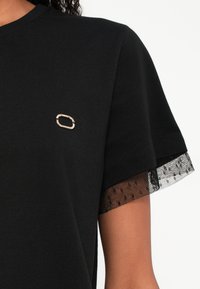 Črna majica s kratkimi rokavi, ki ima prosojni črni čipkasti rob, vezen zlat napis na prsih, gladka bombažna tkanina.