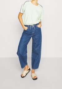 Camiseta verde claro de manga corta con ribetes negros; vaqueros de denim azul sueltos con bolsillos frontales visibles; sandalias beige de tiras cruzadas.