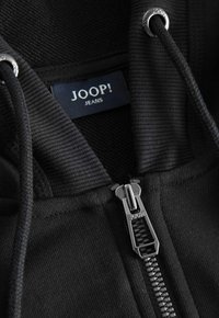 Sudadera con cremallera negra, con textura acanalada, cremallera plateada y etiqueta de logo que dice "JOOP! JEANS" en el área del collar.