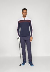 Marineblå golfgenser med glidelås og hvite og mørkerøde detaljer, kombinert med marineblå golfbukser og blå sko, holder en golfkølle.