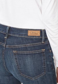 Mörkblå denimjeans med gyllene sömmar, med en loggad läderpatch vid midjan och klassiska bakfickor.