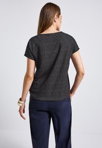Krátký rukáv top v černé tkanině s geometrickým vzorem. Kulatý výstřih, volný střih a hladká textura. Nosit s námořnickými kalhotami.