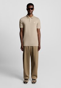 Beige Polohemd mit Struktur und Kragen, kombiniert mit lockeren beigen Hosen. Braune gemusterte Schuhe und dunkle Sonnenbrillen runden das Outfit ab.