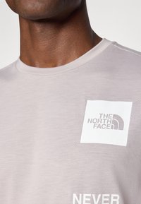 Lysegrå t-skjorte laget av mykt stoff, med et firkantet logomerke med "THE NORTH FACE" i svart og hvit tekst. Rund hals, korte ermer.