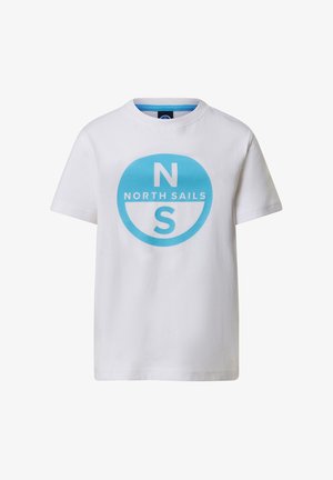 Jeune garçon debout les mains dans les poches portant un t-shirt blanc North Sails, un short bleu clair, des chaussettes blanches et des baskets blanches.