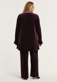 Vrouw staat met haar rug naar ons toe, gekleed in een donkerpaarse lange mouw tuniek en bijpassende losse broek met zwarte hakken.