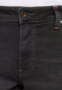 Jeans de mezclilla negra con acabados de costura naranja. Cuenta con un cierre de botón y un diseño estándar de cinco bolsillos. Contiene un detalle de tejido texturizado.