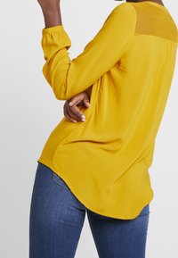 Blusa senza maniche color senape con schiena texturizzata, tessuto morbido, vestibilità ampia e orlo curvo. Abbinata a jeans blu scuro.