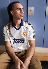 Maillot blanc adidas avec col bleu et accents jaunes, arborant le logo 'Teka' et le blason du Real Madrid. Porté avec un pantalon beige, assis contre des casiers.