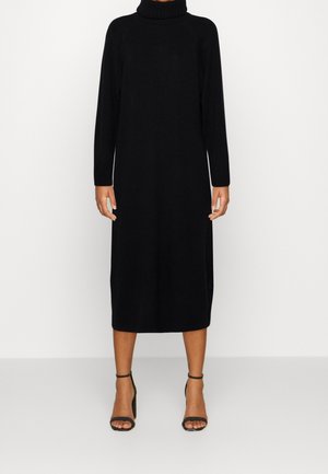 Personne portant une robe midi noire à manches longues et col roulé, avec des sandales à talons hauts noires à bride cheville, debout sur un fond clair uni.
