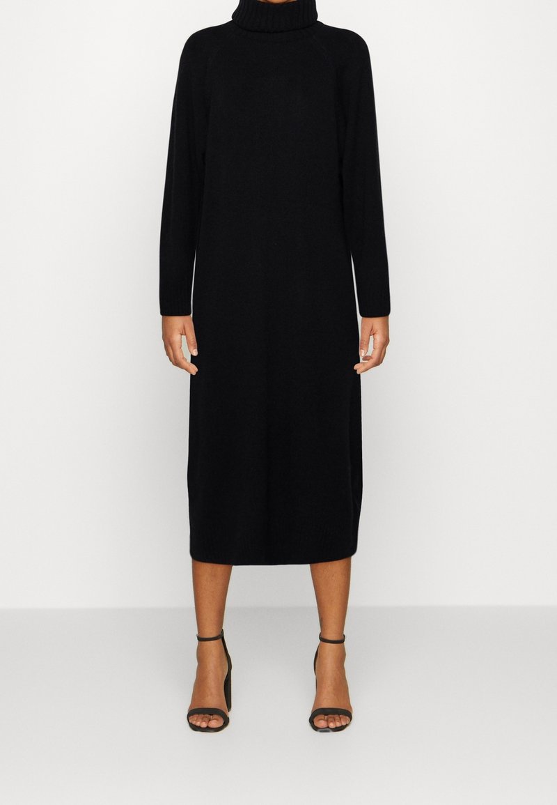 Personne portant une robe midi noire à manches longues et col roulé, avec des sandales à talons hauts noires à bride cheville, debout sur un fond clair uni.
