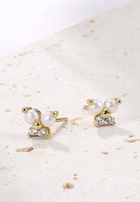 Boucles d'oreilles en or en forme de papillon avec deux perles blanches et deux strass transparents, posées sur une surface texturée de couleur claire.