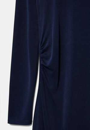 Temno modra majica z dolgimi rokavi s podrobnostjo nabiranja na strani iz gladke tkanine na enobarvnem ozadju.