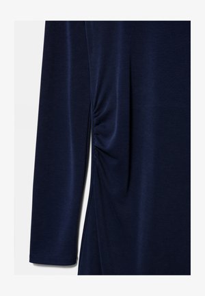 Temno modra majica z dolgimi rokavi s podrobnostjo nabiranja na strani iz gladke tkanine na enobarvnem ozadju.