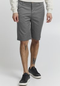 Pantalones cortos de algodón grises con corte recto, que cuentan con bolsillos laterales y un cierre de botón único. Llevados con zapatillas negras.