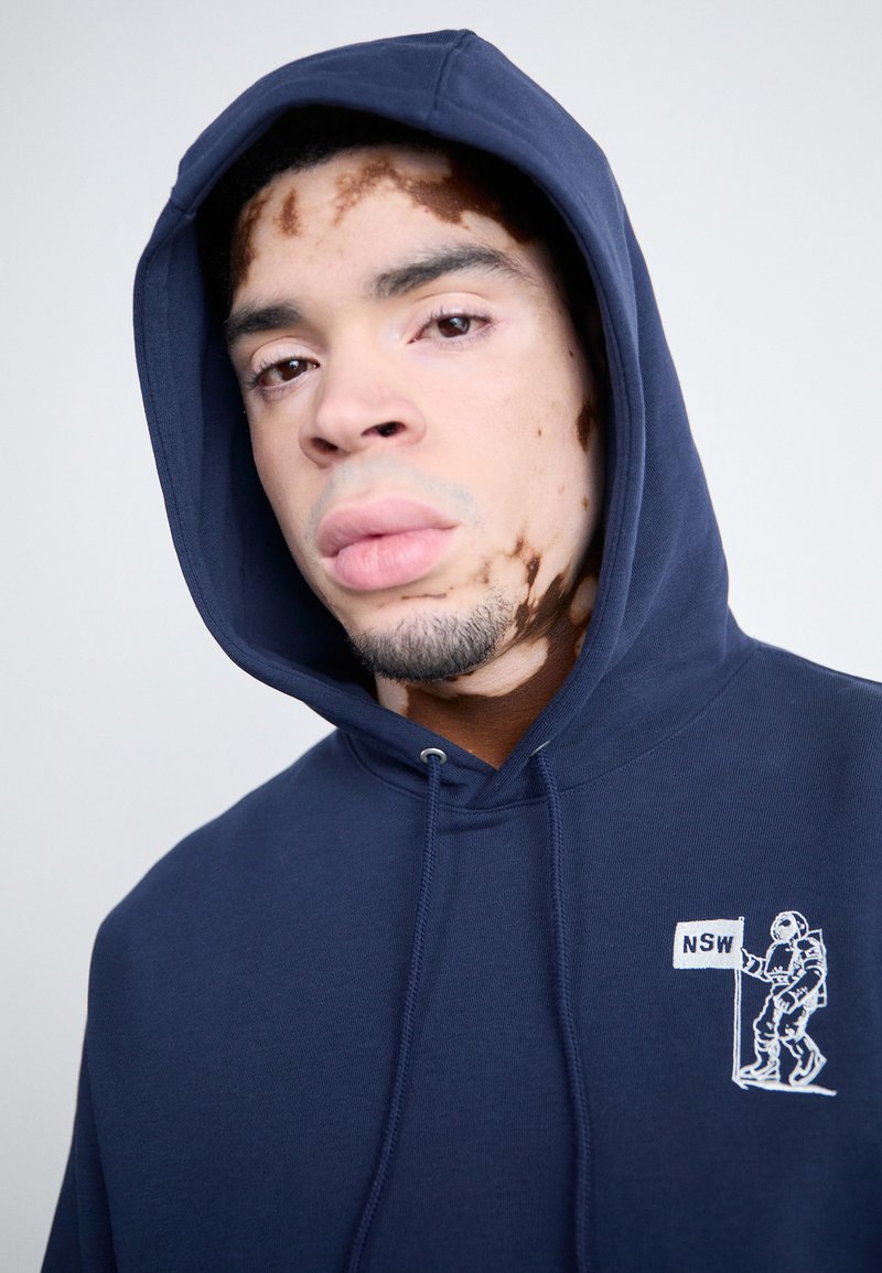 Sudadera azul marino con cordón, que presenta un gráfico bordado de un astronauta sosteniendo una bandera con "NSW" en el pecho. Textura suave.