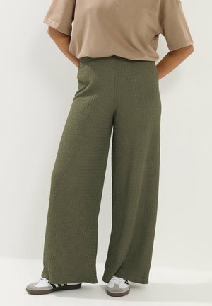 Osoba nosząca teksturowane oliwkowozielone spodnie z szerokimi nogawkami, beżową bluzkę z krótkim rękawem i białe trampki z czarnymi paskami, stojąca z rękami za plecami.