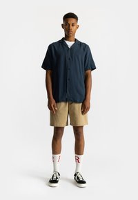 Korte mouwen marineblauw shirt met een knoopsluiting, gecombineerd met beige shorts. Witte sokken met rode opdruk en zwarte sneakers maken de look af.