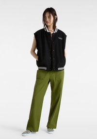 Svart ärmlös varsityjacka med vita ränder, kombinerad med olivgröna vida byxor. Vita och svarta rutiga skor fulländar stilen.